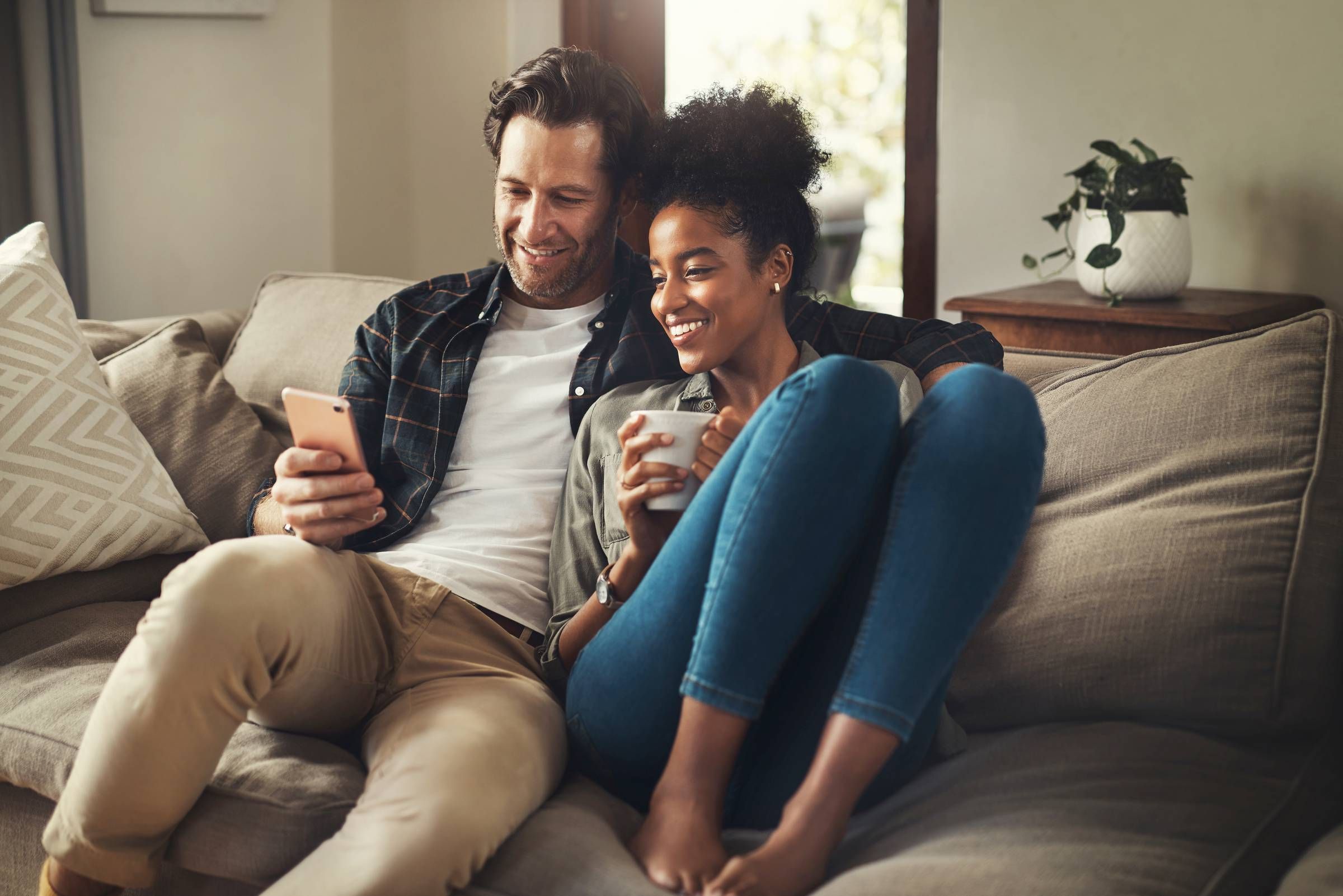 Smiling couple sitting on a couch, looking at a smartphone, with a mug and pillow nearby.