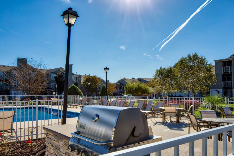 Outdoor pool area with lounge chairs, tables, and a grill under a clear, sunny sky.