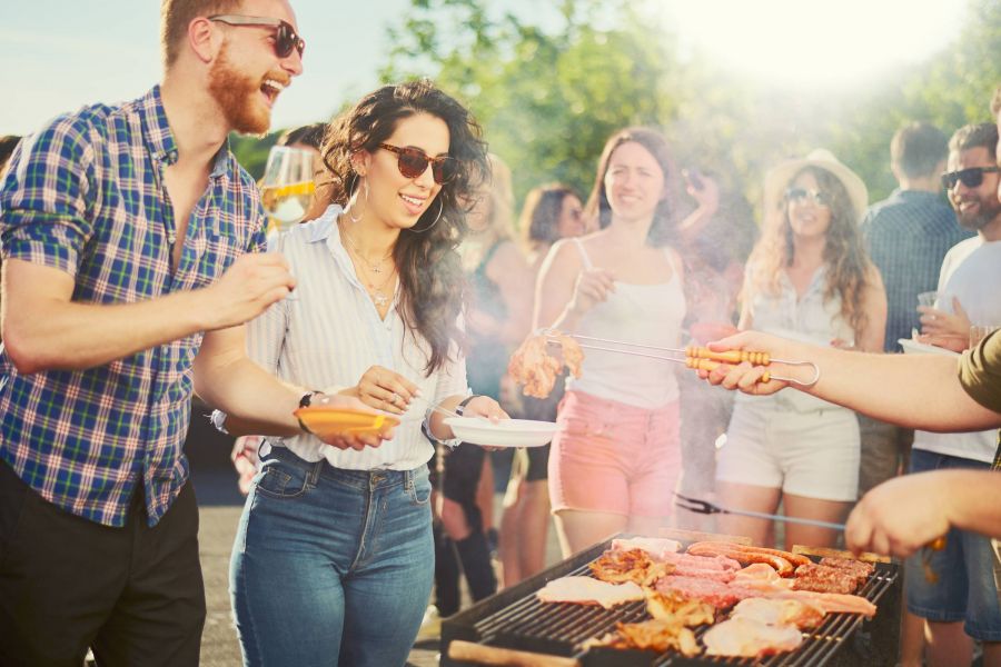 People enjoying a sunny outdoor barbecue, serving food and laughing together with drinks in hand.