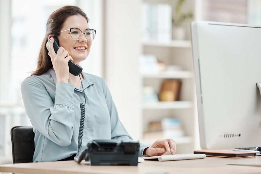 Smiling woman in glasses talks on a corded phone while using a computer at her desk in an office.