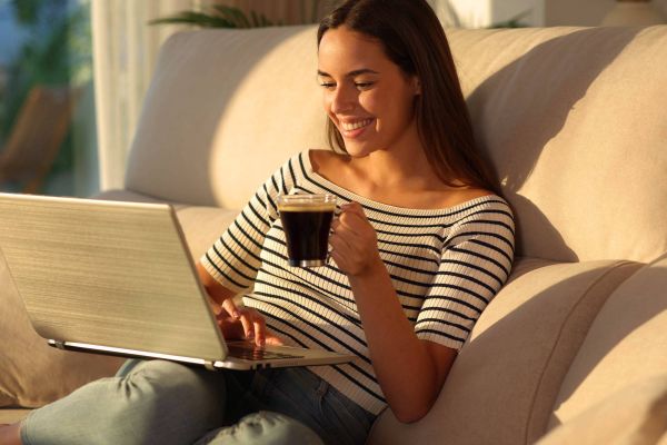 Woman sitting on a couch, smiling at a laptop, holding a coffee mug in a sunlit living room.