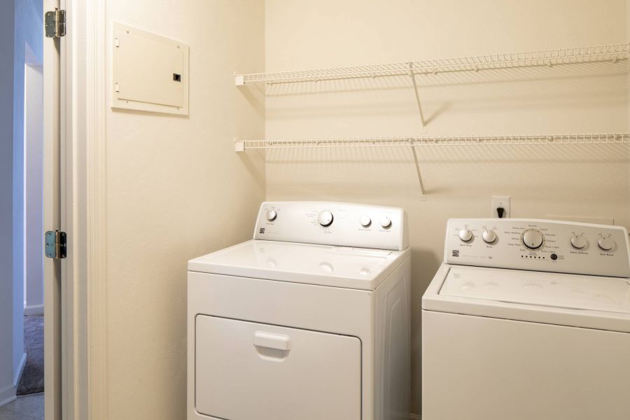 Laundry room with a white washer and dryer, beige walls, and two empty wire shelves above the machines.