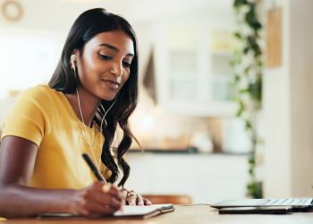 Woman in a yellow shirt writing in a notebook while using a laptop and wearing earphones at a table.