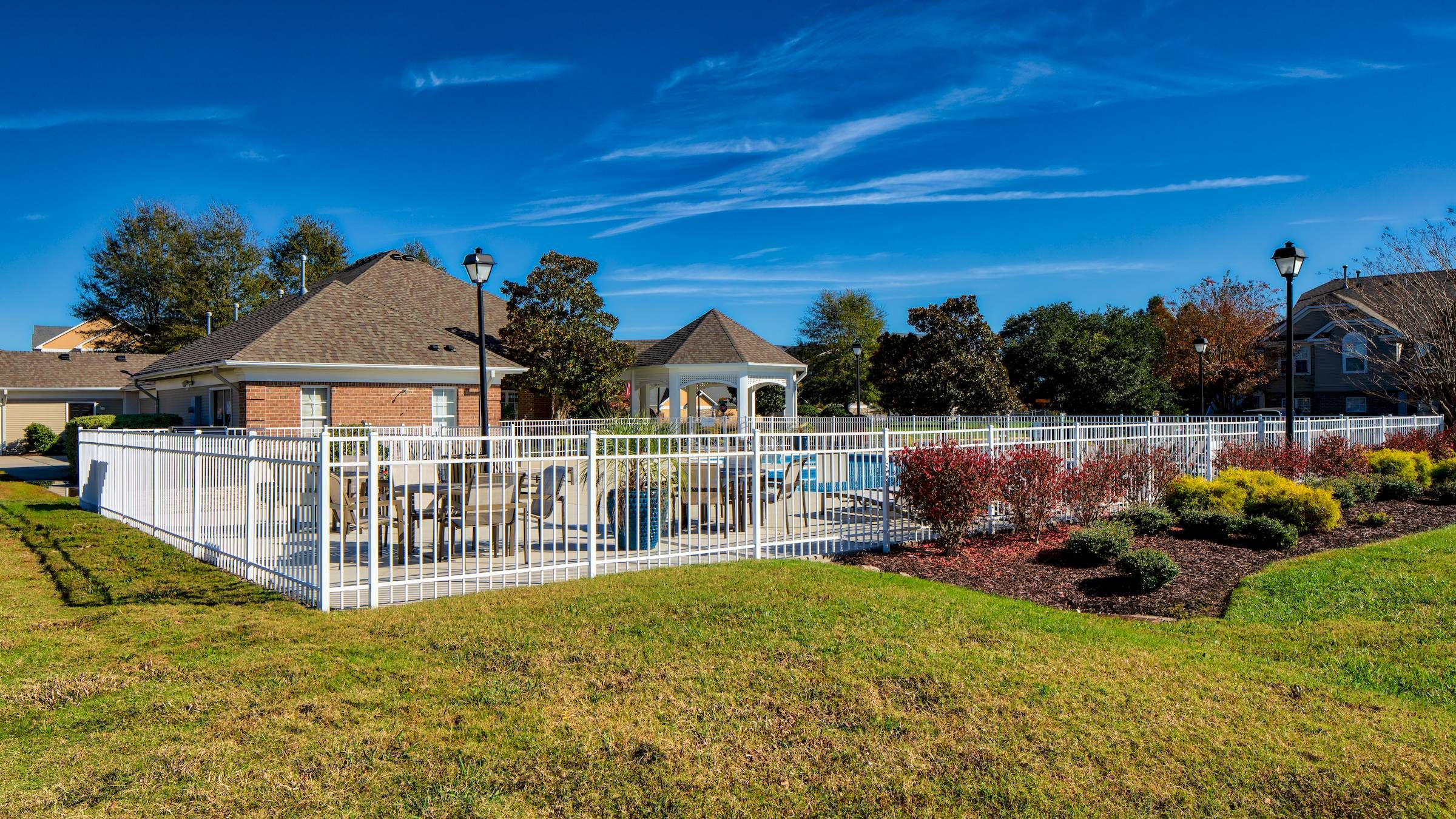Fenced outdoor pool area with lounge chairs, a gazebo, and landscaped garden under a clear blue sky.