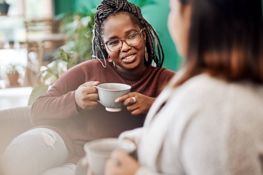 Two women sit and talk, each holding a mug, in a cozy indoor setting with plants in the background.