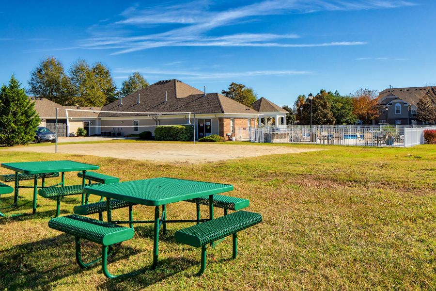 Green picnic tables on a grassy area near a volleyball court, pool, and buildings under a blue sky.