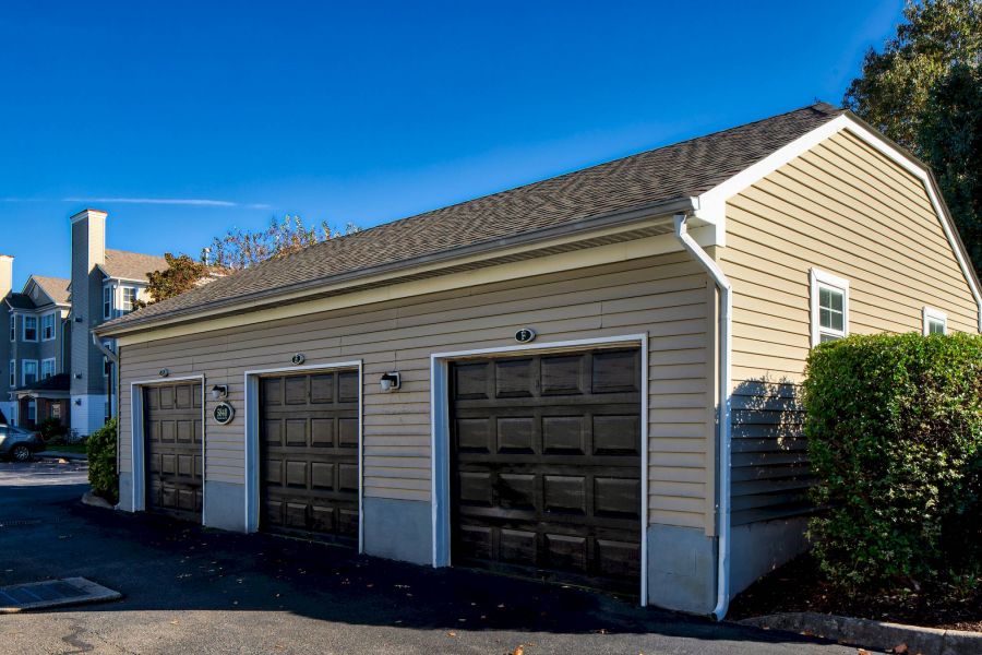 A beige, three-car garage with dark doors, next to bushes and a parking lot under a clear blue sky.