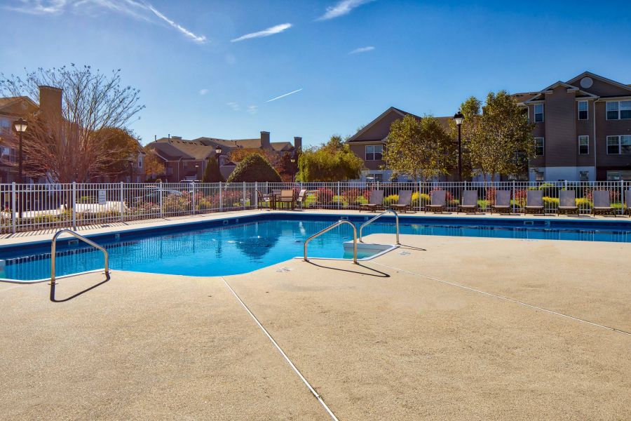 Outdoor swimming pool with metal handrails, surrounded by a fence and apartment buildings under a clear blue sky.