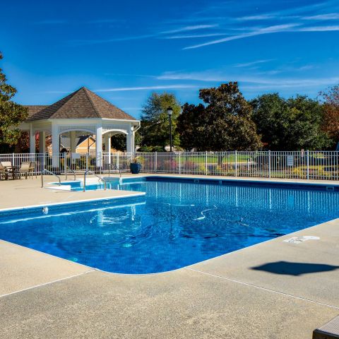 Outdoor swimming pool with a white gazebo, lounge chairs, and buildings in the background on a sunny day.