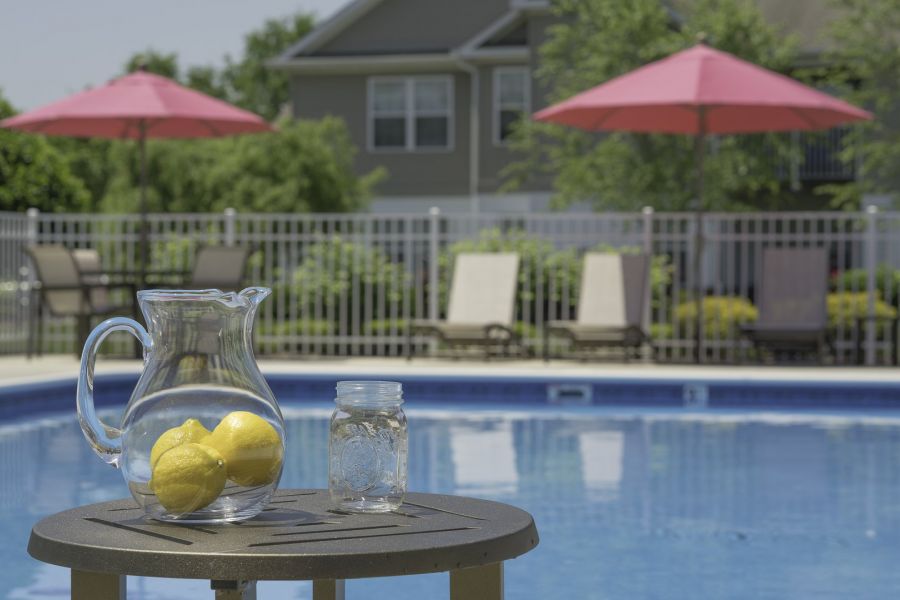 A pitcher with lemons and a glass jar sit on a table by a pool with pink umbrellas and lounge chairs.