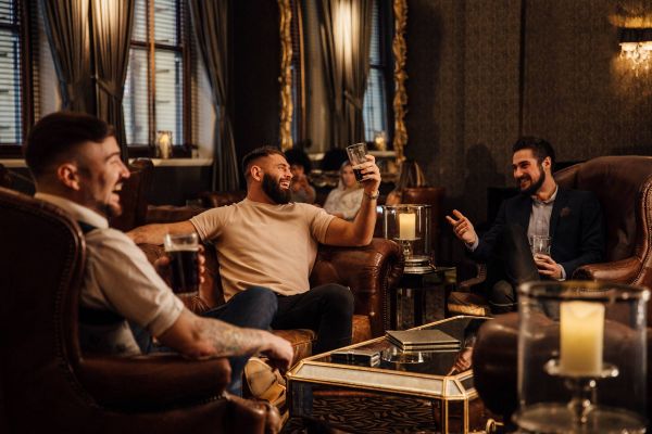 Three men sit in armchairs, laughing and holding drinks in a cozy, dimly lit lounge with candles and mirrors.