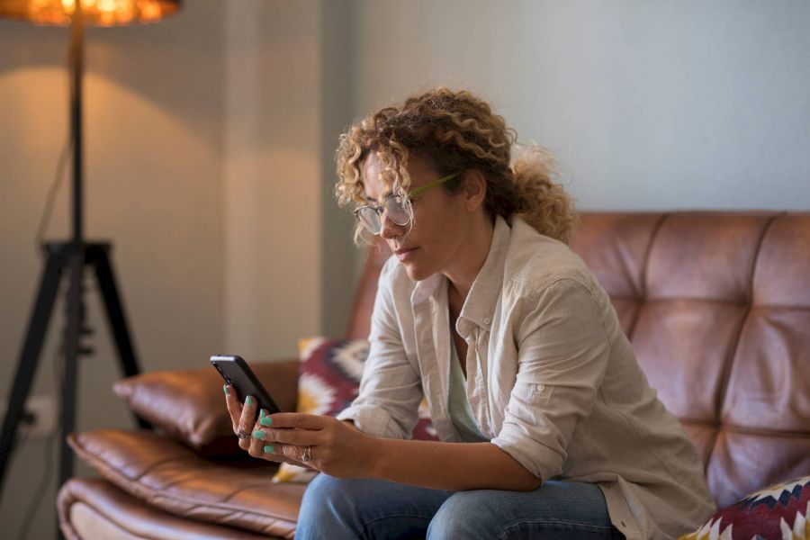 Woman with curly hair and glasses sitting on a couch, looking at her smartphone in a cozy living room.