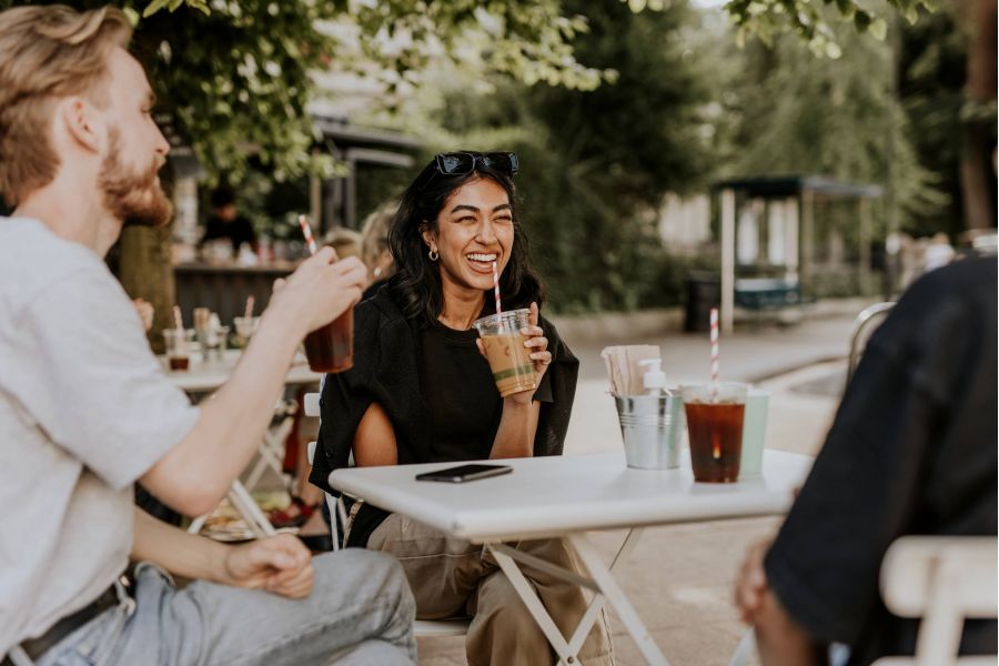 Three people sit at an outdoor café table, laughing and enjoying iced drinks on a sunny day.