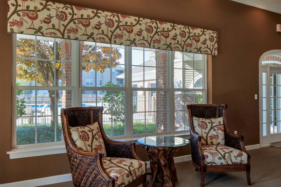 Two armchairs and a round table sit by large windows with floral valances in a sunlit room.