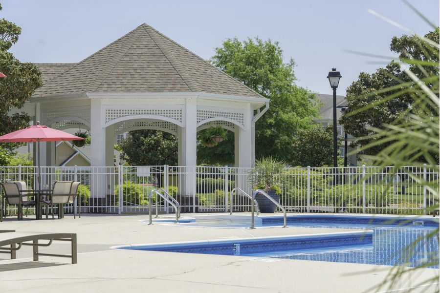 Outdoor pool area with lounge chairs, tables, and a white gazebo surrounded by greenery and a fence.