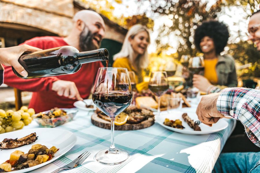 A group of friends enjoys a meal outdoors as wine is poured into a glass on a picnic table.