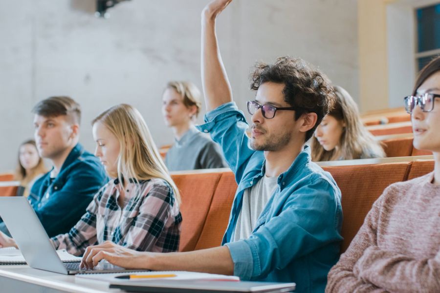 Students in a lecture hall; one student raises his hand while others listen or take notes.
