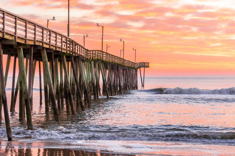 Wooden pier extends over gentle ocean waves at sunrise with a colorful pink and orange sky.