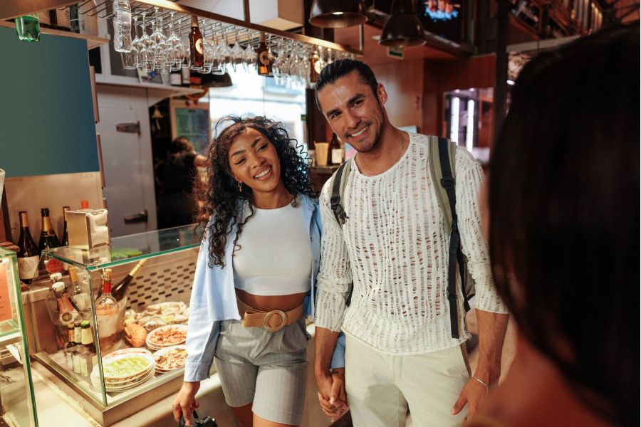 A smiling couple holding hands stands in front of a food display at a market or café.