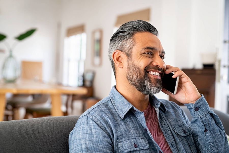 Smiling man with salt-and-pepper hair talks on a smartphone while sitting on a couch at home.
