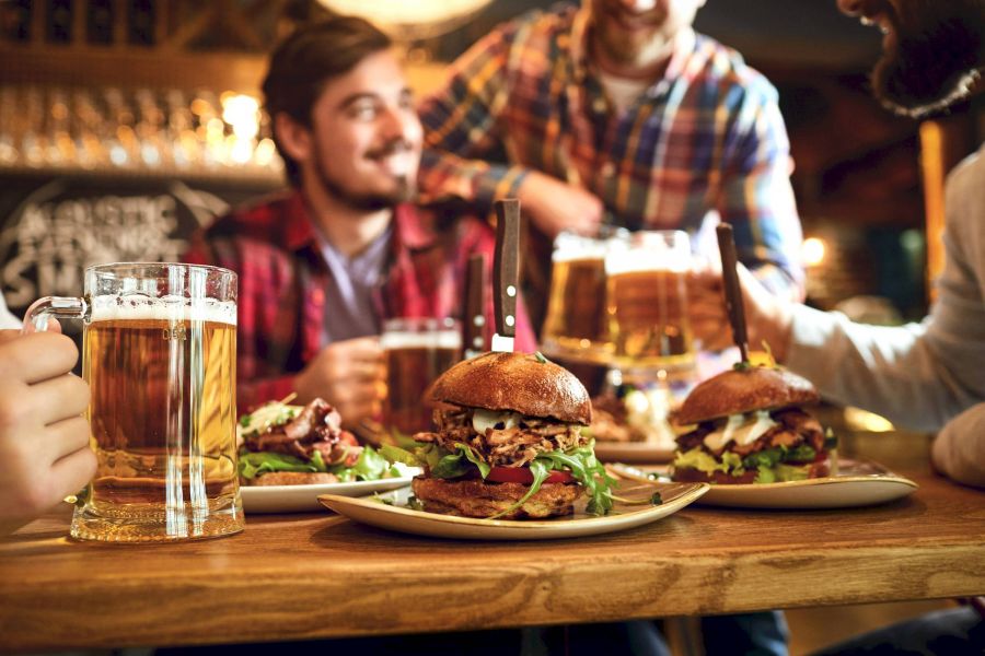 Three people enjoy burgers and beer at a wooden table in a lively, warmly lit restaurant or bar.