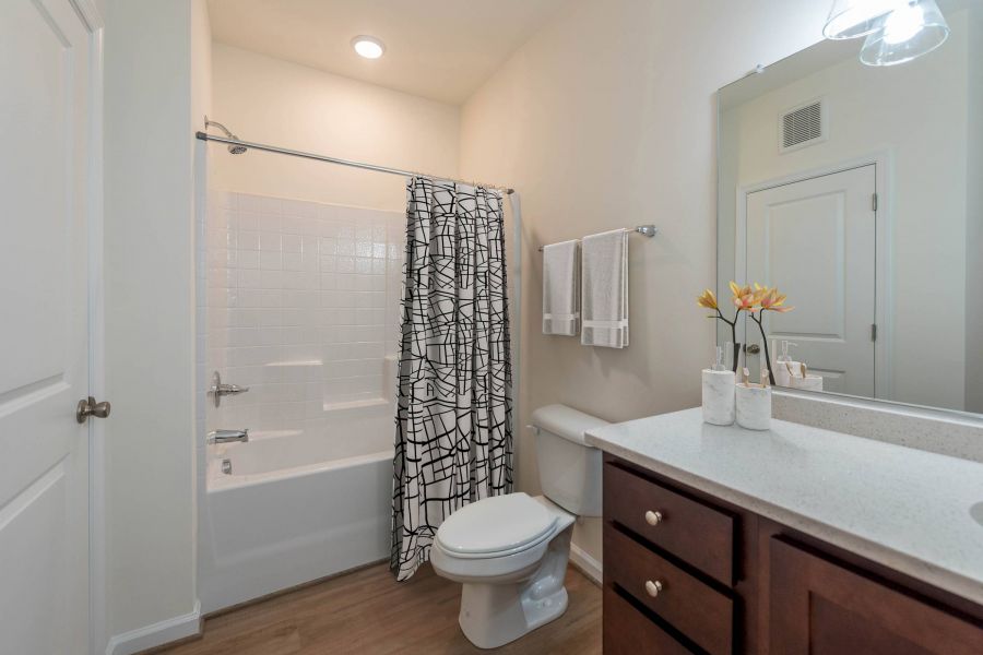 Modern bathroom with a bathtub, geometric shower curtain, toilet, and vanity with a mirror and flowers.